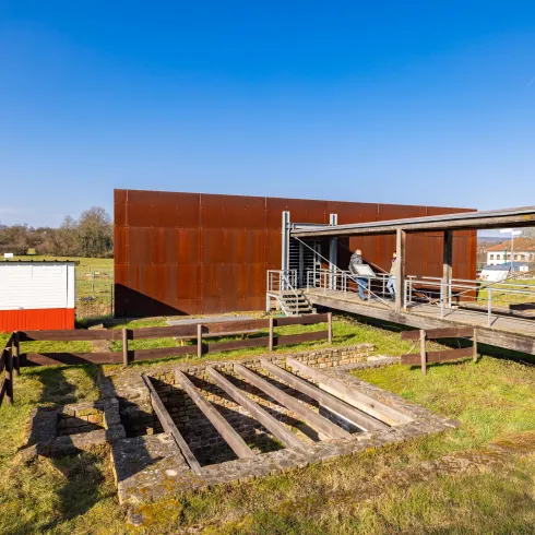Ein modernes Gebäude mit einer rostigen Metallfassade steht in einer grünen Landschaft. Im Vordergrund sind Überreste einer alten Struktur zu sehen.