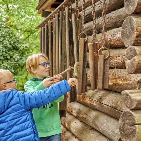 Zwei Kinder spielen an einer Holzwand und erkunden Klanginstallationen. Sie zeigen neugierig auf die hängenden Metallröhren.