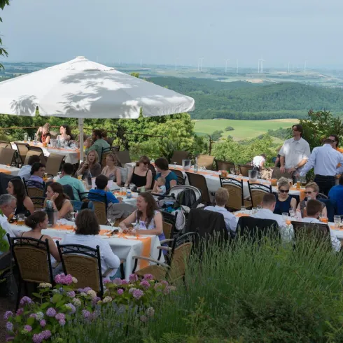 Eine große Außenterrasse mit vielen Gästen, die an Tischen sitzen. Im Hintergrund ist eine schöne Landschaft mit Hügeln und Bäumen zu sehen.