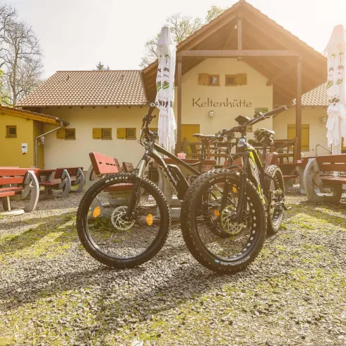 Zwei Fahrräder stehen vor einem gemütlichen Häuschen mit Terrassenmöbeln. Im Hintergrund sind Bäume und Sonnenschein zu sehen, was eine angenehme Atmosphäre schafft.