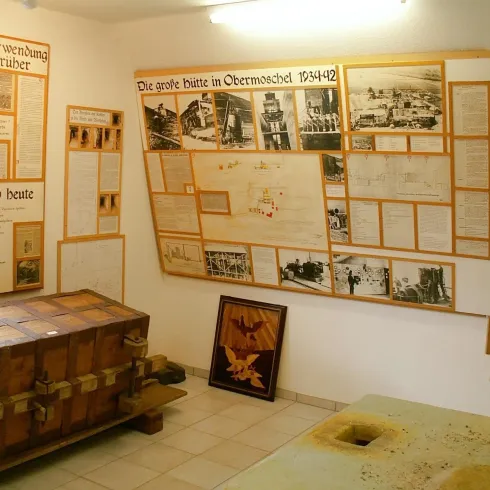 Ein Raum mit historischen Ausstellungen, der Informationen über die Ereignisse von 1944 in Obermoschel präsentiert. An der Wand hängen Dokumente und Bilder, während ein Holzkasten im Vordergrund steht.
