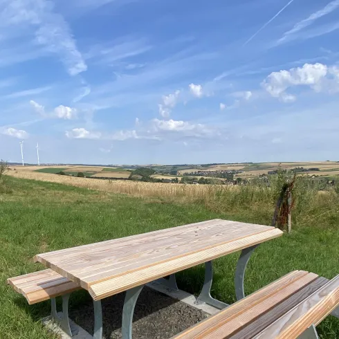 Ein Holzpicknicktisch steht in einer grünen Wiese mit Blick auf Felder und einen blauen Himmel. Sanfte Hügel und einige Windräder sind im Hintergrund sichtbar.