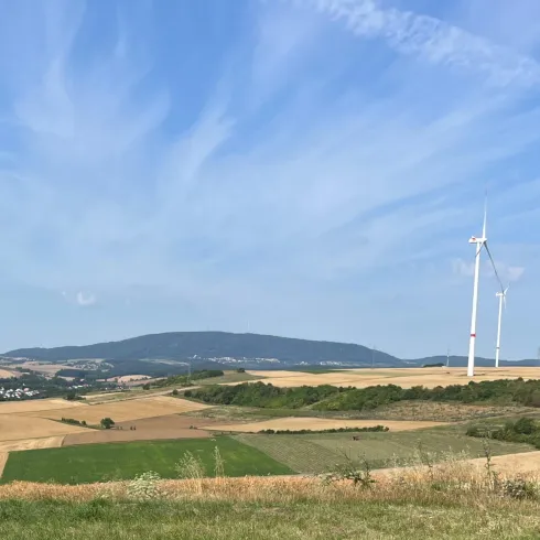Eine weite Landschaft mit sanften Hügeln und Feldern. Im Hintergrund sind mehrere Windkraftanlagen und ein blauer Himmel mit leichten Wolken.