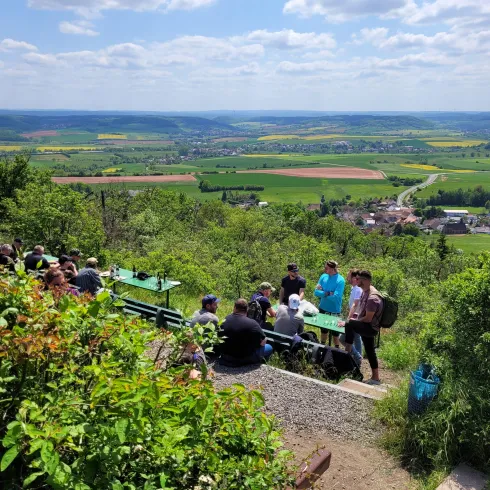 Eine Gruppe von Menschen sitzt an einer Aussichtsterrasse mit Blick auf grüne Felder und Hügel. Der Himmel ist blau mit einigen Wolken.