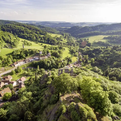 Eine wunderschöne Landschaft mit grünen Hügeln und bewaldeten Bereichen. Im Vordergrund sind kleine Häuser zu sehen, die sich harmonisch in die Natur einfügen.