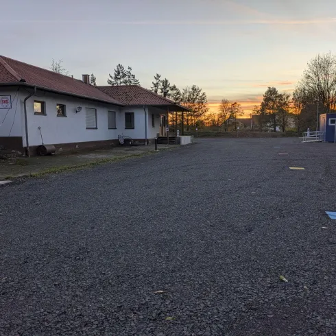 Ein ruhiger Parkplatz vor einem Gebäude bei Sonnenuntergang. Die Umgebung ist ruhig mit Bäumen im Hintergrund.