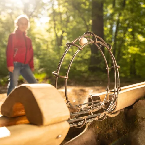 Ein Spielgerät im Wald aus Holz und Metall. Im Hintergrund ist ein Kind in einem roten Kleidungsstück zu sehen.