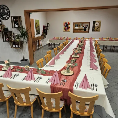 Ein festlich gedeckter Tisch in einem Restaurant mit weißen Tischdecken und rosa Servietten. Die Stühle sind aus Holz und es gibt dekorative Elemente auf dem Tisch.