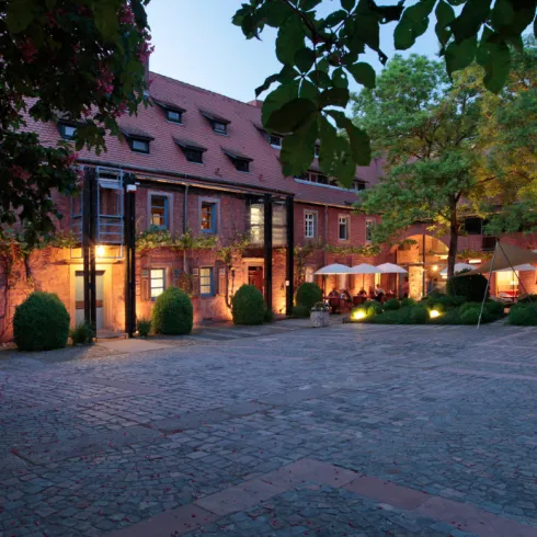 Eine idyllische Hofstraße mit historischen Gebäuden und sanfter Beleuchtung. Grüne Bäume und gemütliche Sitzgelegenheiten schaffen eine einladende Atmosphäre.