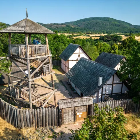 Eine ländliche Szene mit einem Aussichtsturm und traditionellen Fachwerkhäusern. Umgeben von Bäumen und sanften Hügeln bietet der Ort eine ruhige Atmosphäre.