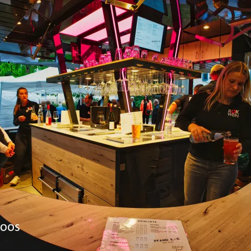 Eine moderne Bar mit einer Person, die ein Getränk einschenkt. Im Hintergrund sind Gäste und eine einladende Atmosphäre zu sehen.