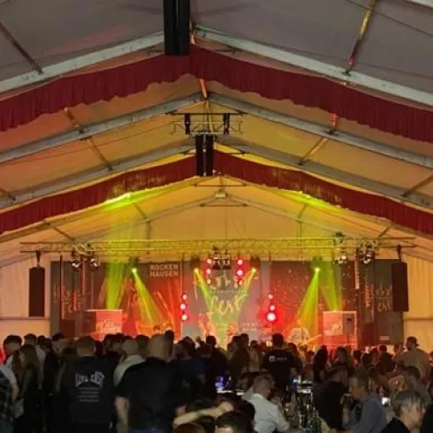 Ein überfülltes Zelt mit Musikern auf der Bühne und bunten Lichtern. Die Atmosphäre ist festlich und lebhaft, mit vielen Menschen, die feiern.