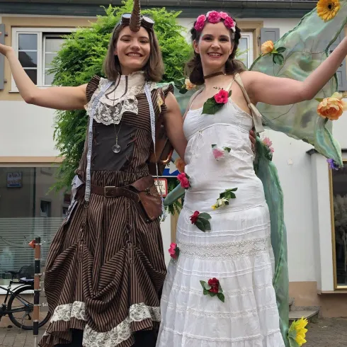Zwei junge Frauen in fantasievollen Kostümen stehen fröhlich nebeneinander. Die eine trägt ein elegantes weißes Kleid mit Blumen, die andere ein braunes, gestreiftes Outfit mit einem Einhorn-Horn.