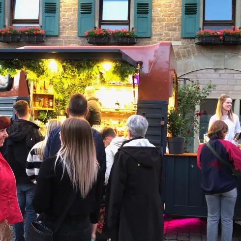 Ein lebhaftes Straßenfest mit einem mobilen Verkaufsstand und vielen Menschen. Die Atmosphäre ist freundlich und einladend.