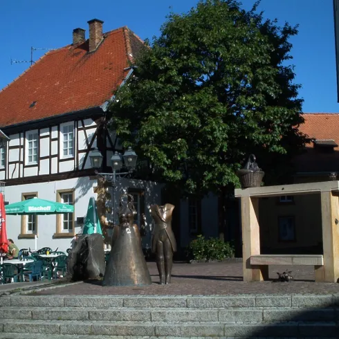 Ein schöner Platz mit einer Bronzeskulptur und einem großen Baum. Im Hintergrund sind traditionelle Gebäude und Sitzgelegenheiten im Freien sichtbar.