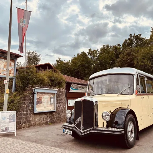 Ein alter Bus steht vor einem Informationszentrum in einer grünen Umgebung. Der Himmel ist bewölkt und es gibt einige Schilder in der Nähe.