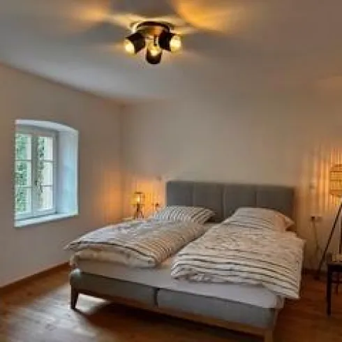 Ein gemütliches Schlafzimmer mit einem Doppelbett und sanftem Licht. Die Wände sind hell und ein Fenster sorgt für Tageslicht.