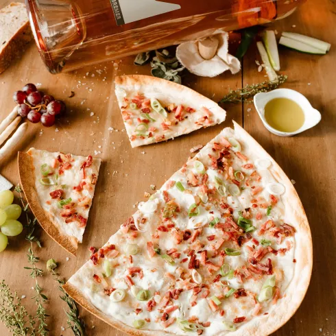 Eine köstliche Pizza mit Schinken und Gemüse liegt auf einem Holztisch. Daneben steht eine Flasche Roséwein und frische Zutaten.