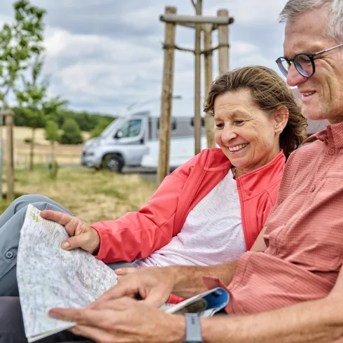 Ein Paar sitzt gemütlich auf einer Bank und schaut auf eine Landkarte. Im Hintergrund ist ein Wohnmobil und eine grüne Landschaft zu sehen.