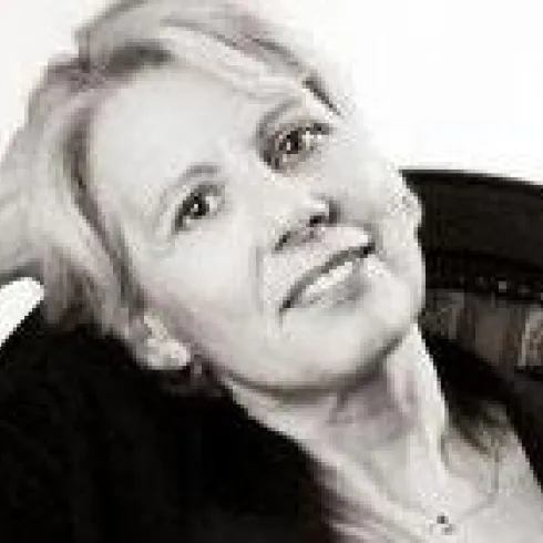 Eine Frau mit blonden Haaren sitzt entspannt auf einem Stuhl. Sie lächelt freundlich in die Kamera.