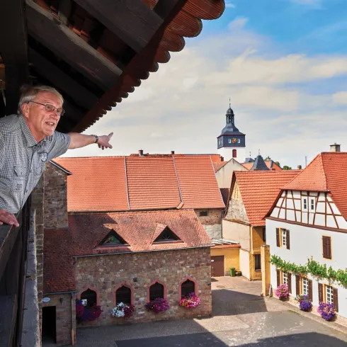 Ein älterer Mann steht auf einem Balkon und zeigt auf eine malerische Dorfansicht. Die Gebäude haben traditionelle Architektur mit roten Dächern und einer schönen Wolkenlandschaft im Hintergrund.