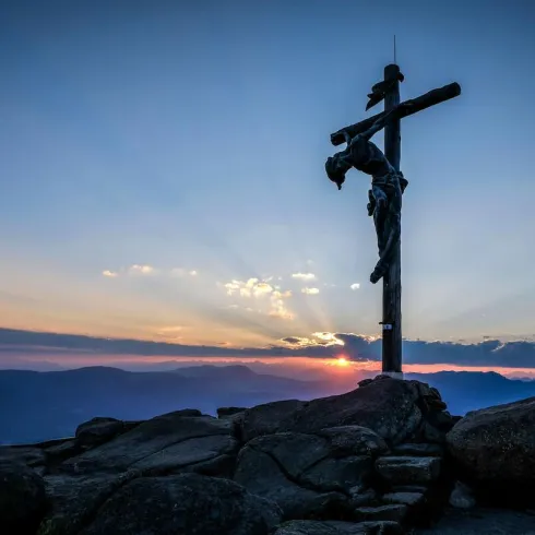Ein Kreuz steht auf einem Berggipfel vor einem farbenfrohen Sonnenuntergang. Die Landschaft im Hintergrund erhebt sich mit sanften Hügeln und Wolken.