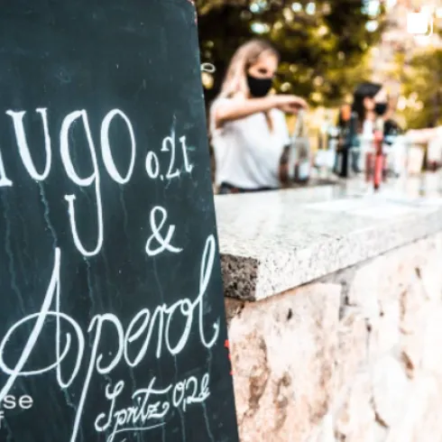 Ein schwarzes Schild mit der Aufschrift "Hugo 0,2 l & Aperol" steht an einer Bar. Im Hintergrund sieht man eine Person, die Getränke zubereitet.