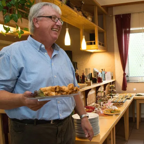Ein lächelnder Mann hält einen Teller mit Essen in einer Einrichtung. Im Hintergrund ist ein Tisch mit verschiedenen Speisen und Getränken sichtbar.