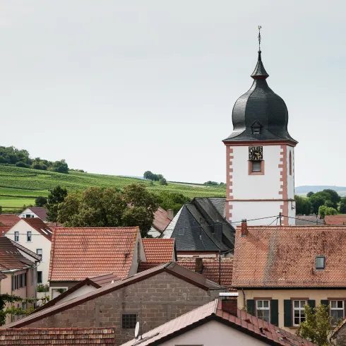 Eine malerische Dorfansicht mit einer markanten Kirchturm. Im Hintergrund sind sanfte Hügel und Windkraftanlagen zu sehen.