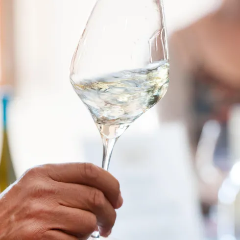 Ein Mann hält ein Weinglas mit hellem Wein in der Hand. Im Hintergrund sind verschwommene Silhouetten von anderen Menschen sichtbar.